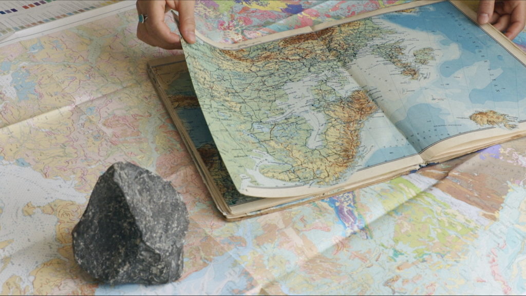 Hand flipping a page of an atlas with a stone upon a flat map in the foreground.