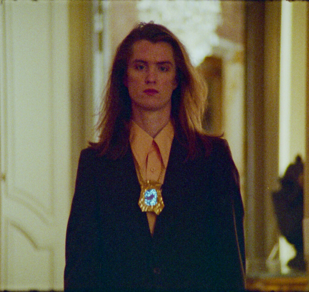 A person strolls through a long hallway in front of a crystal chandelier, wearing a glowing brass video player necklace, a smart jacket and on oversized collared shirt with long blonde hair flowing.